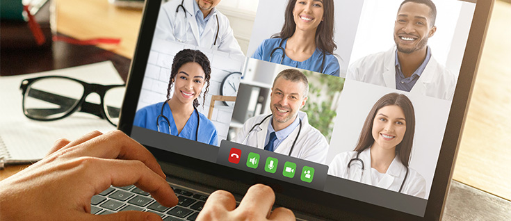 A pair of hands typing on a laptop with the screen showing a telemedicine video call with doctors.
